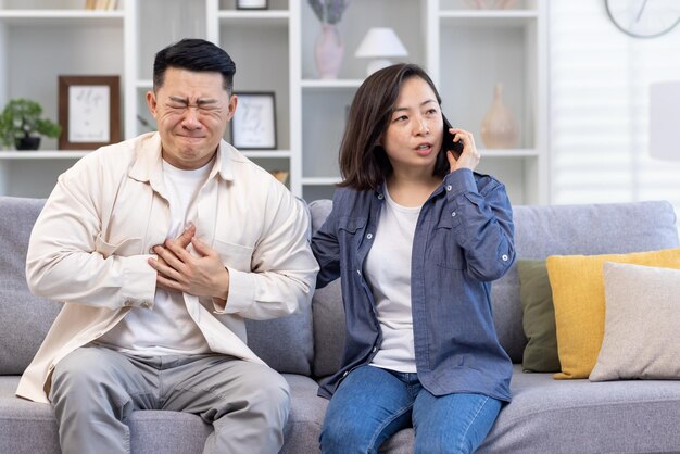 Молодая азиатская семья сидит дома на диване, мужчина прижимает руку к груди, сердечный приступ инсульт