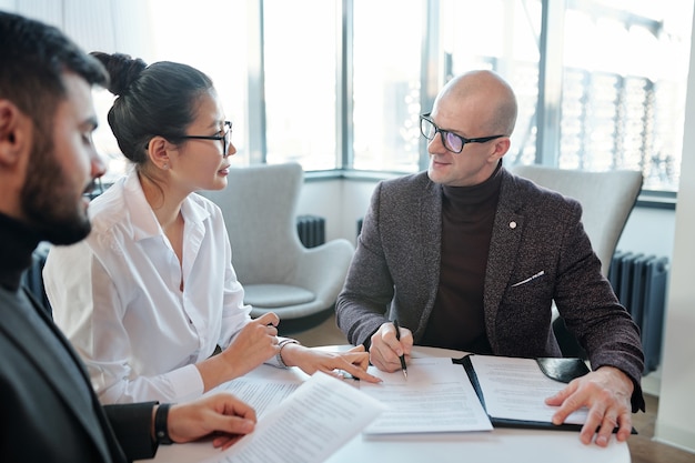 Young Asian female agent looking at mature business partner going to put his signature in contract after negotiation