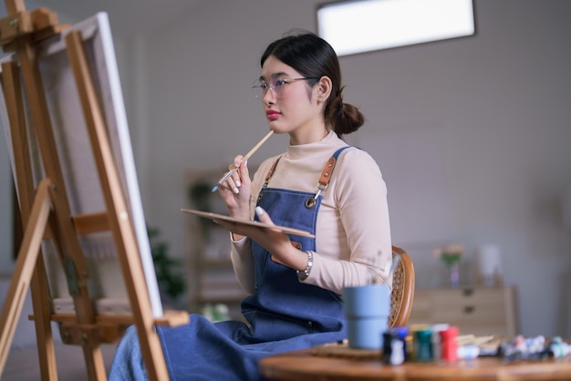 젊은 아시아 여성 예술가 는 캔버스 에서 생각 하는 생각 을 그리는 예술 작품 을 보면서  ⁇ 솔 을 들고 있다