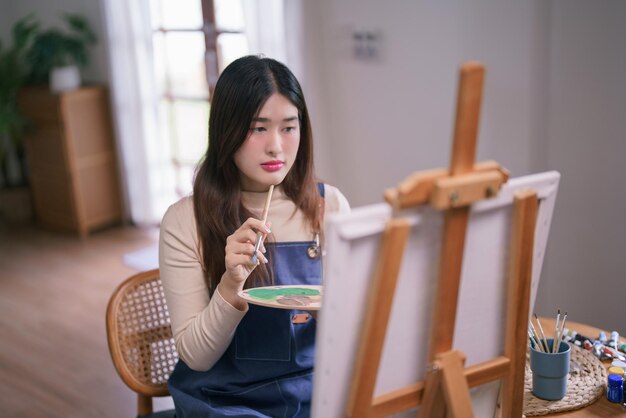 사진 젊은 아시아 여성 예술가가 캔버스 앞에 앉아 그림을 그리고 그림을 그리는 아이디어를 생각하고 있습니다.