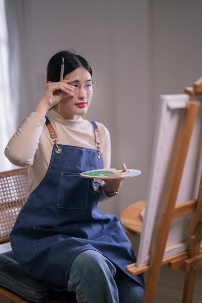 Молодая азиатская художница, касающаяся очков и смотрящая на холст, думает, рисует идеи, рисует произведения искусства