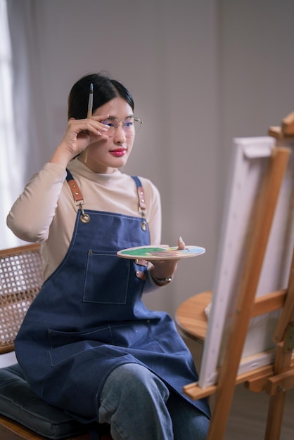 Young asian female artist touching glasses and looking on canvas to thinking idea painting artwork