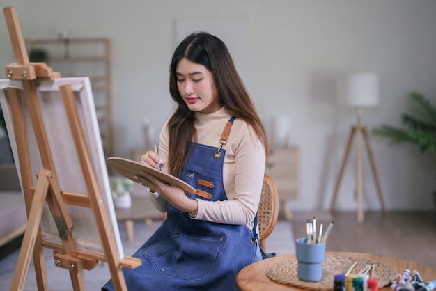 사진 젊은 아시아 여성 예술가 가 캔버스에 그림을 그리기 위해 컬러 패을 가진 페인트브러스를 사용합니다.