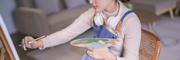 アジアの若い女性アーティストが首にヘッドフォンをかぶってキャンバスにペイントブラシで絵を描いています