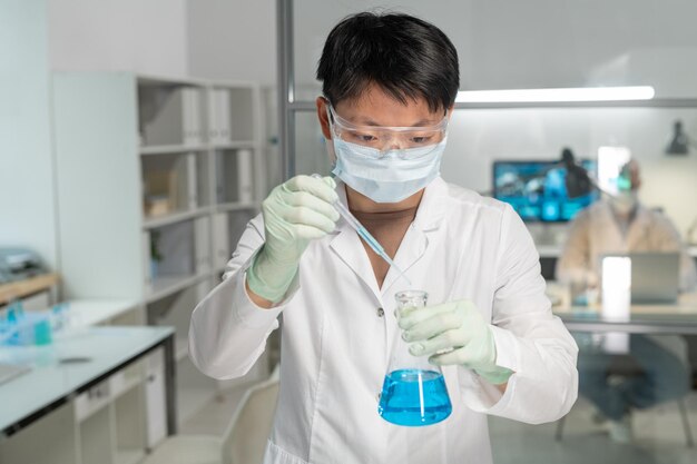 Young asian female chemist adding drop of chemical substance into blue liquid