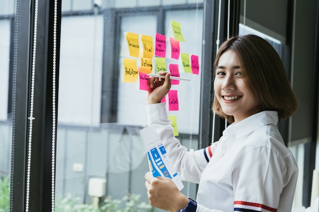 젊은 아시아 여성 창조적 인 모바일 응용 프로그램 개발자 사무실 유리 벽에 다채로운 스티커 메모를 작업.