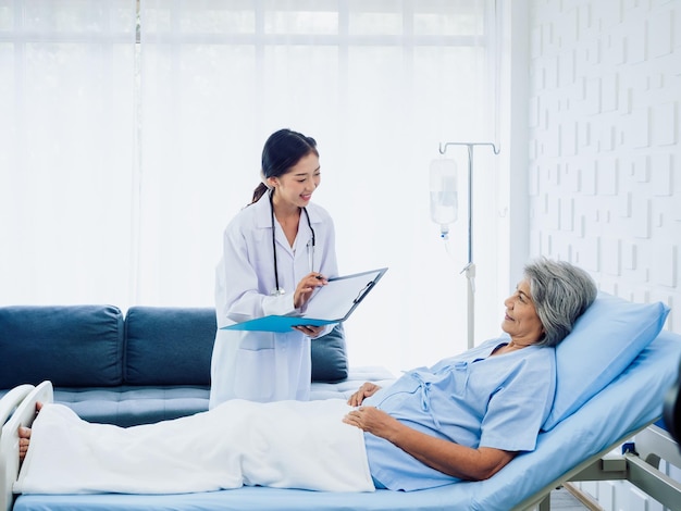 Young Asian female doctors visit senior woman patient in bed explaining the symptoms and examination notes and medical healthcare documents on clipboard Physician in white suit working in hospital