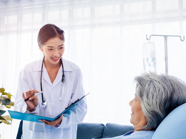 Young Asian female doctors visit senior woman patient in bed explaining the symptoms and examination notes and medical healthcare documents on clipboard Physician in white suit working in hospital