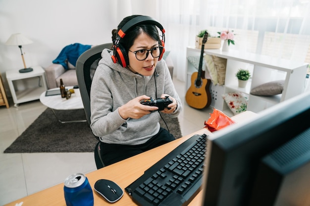 young asian female gamer homebody stay at home on summer break playing online game on computer by controller. concentrated nervous woman nerd in eyeglasses and headset looking on screen internet.