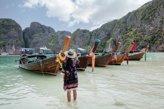 Young asian female raising arm with happiness on coastline with wooden boat in Maya bay at Phi Phi Island, Krabi