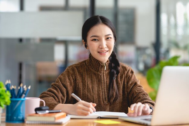 Young Asian female student sitting taking notes and using laptop computer at table Looking at camera