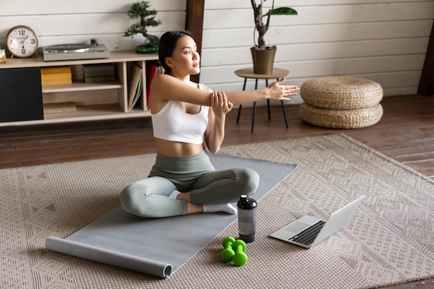 Photo young asian fitness girl doing stretching sport workout at home stretch arms and looking at laptop f...