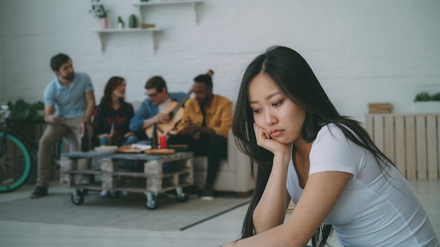 Young asian girl feels upset and isolated while her flatmates\
celebrating party at home indoors