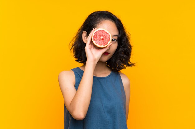 Young asian girl holding a grapefruit over isolated orange background