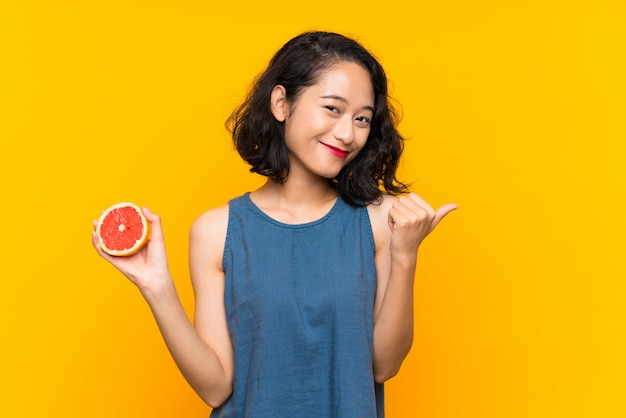 Молодая азиатская девушка держа грейпфрут над изолированной оранжевой стеной указывая к стороне для того чтобы представить продукт