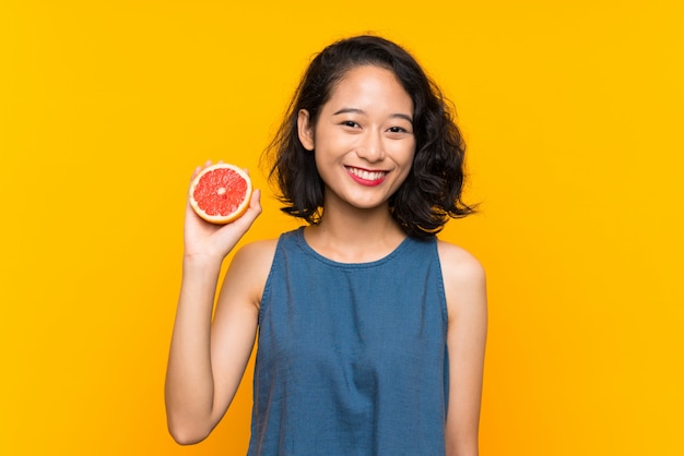 Young asian girl holding a grapefruit over isolated orange wall smiling a lot