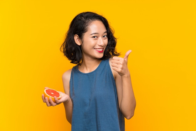 Young asian girl holding a grapefruit over isolated orange wall with thumbs up because something good has happened