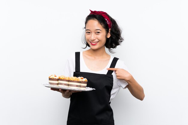 Young asian girl holding lots of muffin cake over isolated white background with surprise facial expression