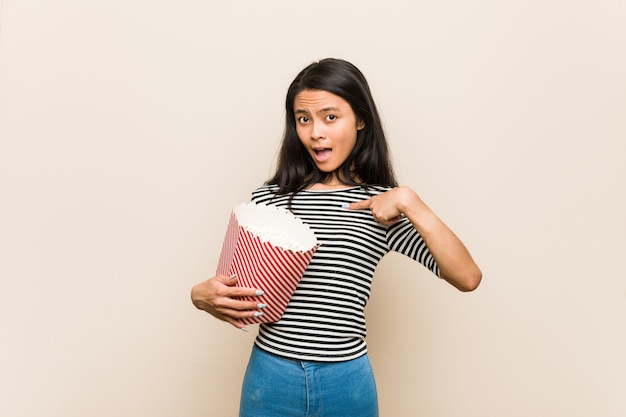 Young asian girl holding a popcorn bucket surprised pointing at himself, smiling broadly.