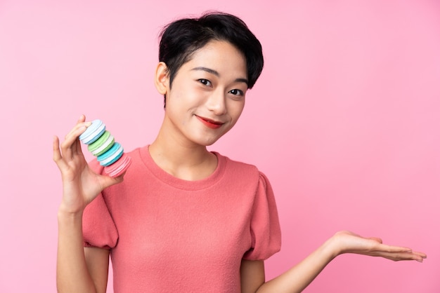 Young Asian girl over isolated pink wall holding colorful French macarons and making doubts gesture