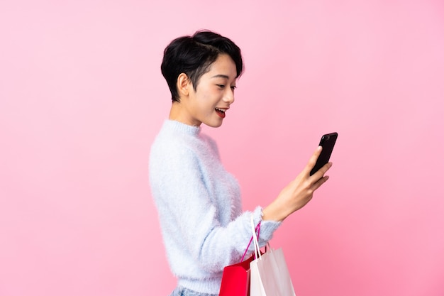 Young Asian girl over isolated pink wall holding shopping bags and writing a message with her cell phone to a friend