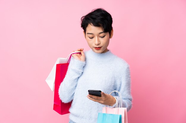 Young Asian girl over isolated pink wall holding shopping bags and writing a message with her cell phone to a friend