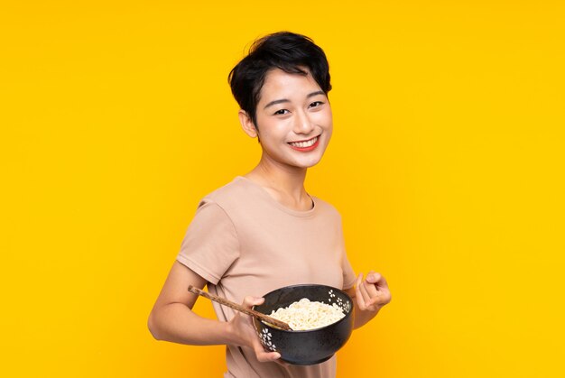 Young Asian girl over isolated yellow wall and pointing it while holding a bowl of noodles with chopsticks