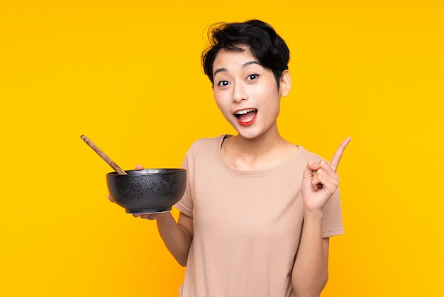 Young Asian girl over isolated yellow wall pointing up a great idea while holding a bowl of noodles with chopsticks