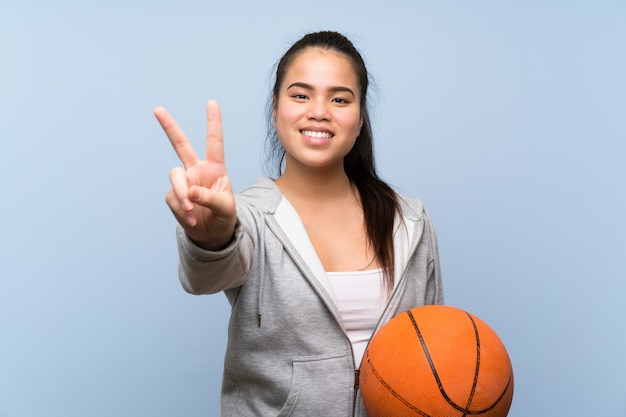 Young Asian girl playing basketball over isolated wall smiling and showing victory sign