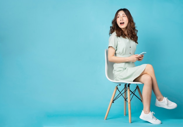 Young Asian girl sitting in chair using phone with surprised expression on turquoise