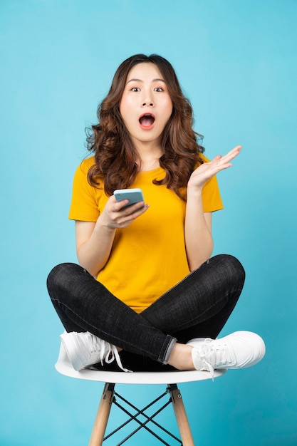 Young asian girl sitting in chair using phone with surprised expression on turquoise