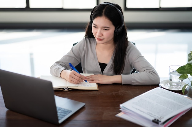 Young asian girl student wears wireless headphones write on the notebook to study language online watch and listen to the lecturer, webinar via video call e-learning at home, distance education