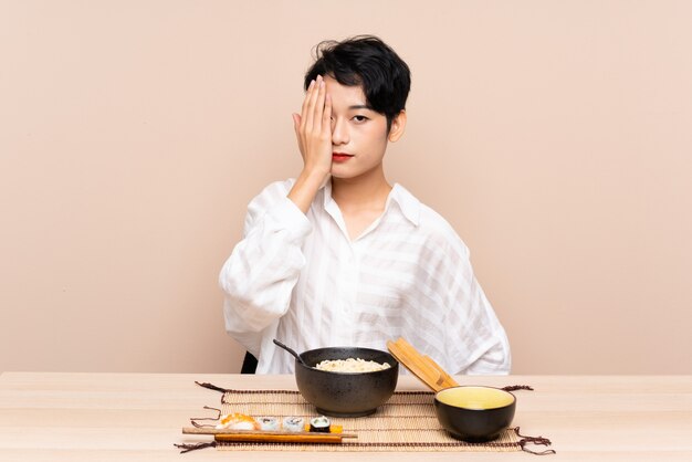 Young Asian girl in a table with bowl of noodles and sushi covering a eye by hand