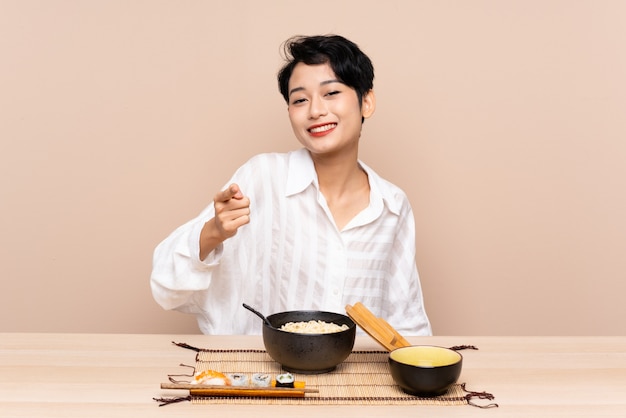 Young Asian girl in a table with bowl of noodles and sushi points finger at you