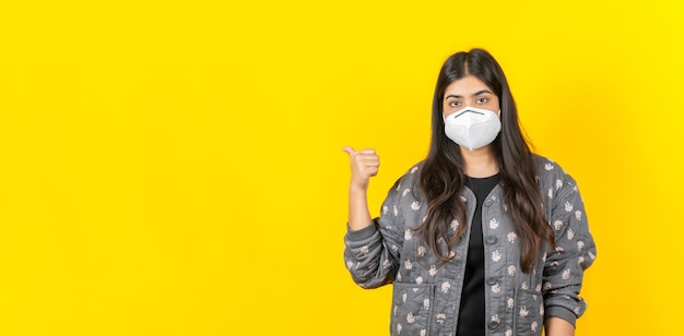 黄色の背景で隔離のカジュアルな服装で医療用フェイスマスクを身に着けている若いアジアの女の子