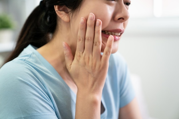 Молодая азиатская девушка с зубной болью и стоматологическими проблемами. японская случайная женщина трогает руками лицо, чувствуя чувствительные зубы внутри. хмурится от несчастного дискомфорта, болезнь жены в помещении в синей футболке.