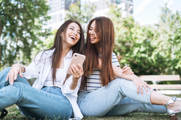 アジアの若い女の子の友達が携帯電話を使って市内の公園で楽しんでいます