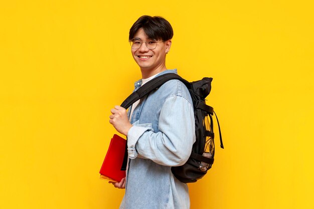 Young asian guy student with backpack and books standing and smiling on yellow isolated background