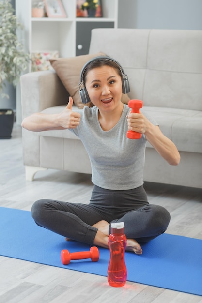 Giovane donna asiatica in buona salute con le cuffie che si esercitano al coperto a casa con una bottiglia d'acqua e manubri in soggiorno seduto sul tappetino da yoga che mostra i pollici in su sorridente