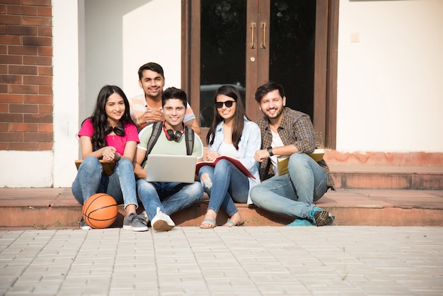 Young Asian Indian college students reading books, studying on laptop, preparing for exam or working on group project while sitting on grass, staircase or steps of college campus