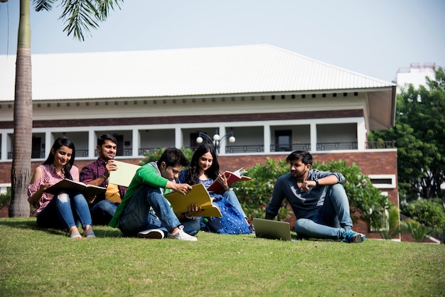 Young asian indian college students reading books, studying on\
laptop, preparing for exam or working on group project while\
sitting on grass, staircase or steps of college campus