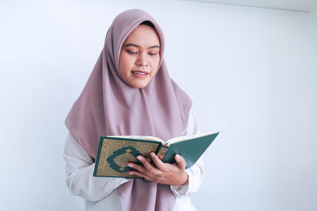 머리 스카프를 두른 젊은 아시아 이슬람 여성이 회색 배경에 미소와 진지한 얼굴로 이슬람의 경전을 기도하거나 읽고 있다