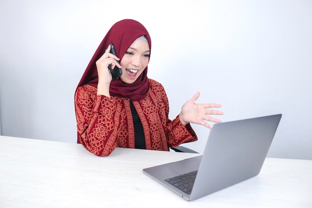 Young asian islam woman wearing headscarf is smiling and\
excited with when she call on the phone and front on the laptop on\
the table
