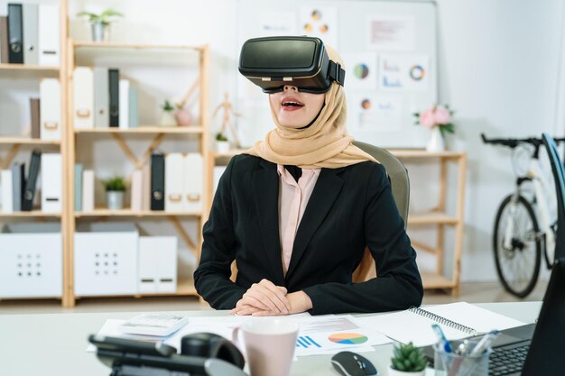 Молодая азиатская исламская работница в профессиональном черном костюме развлекается в очках виртуальной реальности в офисе. женщина-мусульманка сидит за рабочим столом в современной студии и смеется с гарнитурой vr