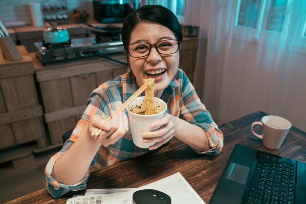 늦은 밤 마감일을 하면서 식당에서 젓가락으로 국수를 먹고 웃고 있는 젊은 아시아 일본 여성. 여자는 집 부엌에서 건강에 해로운 패스트 푸드라면 수프를 즐긴다. 얼굴 카메라 미소