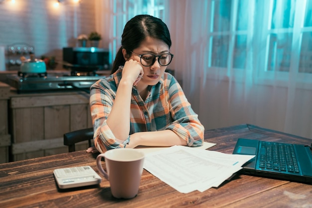 Young asian korean woman in casual wear and glasses getting
stressed over finances in kitchen at midnight. vintage style of
pensive girl sitting and frowning thinking solution with tax
document.