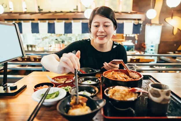 일본 현지 식당에서 반찬을 먹고 스마트폰으로 문자를 보내는 젊은 아시아 한국 여성. 휴대 전화를 사용하고 젓가락을 들고 웃는 소녀는 구운 장어 밥과 함께 쟁반에 전채를 시도