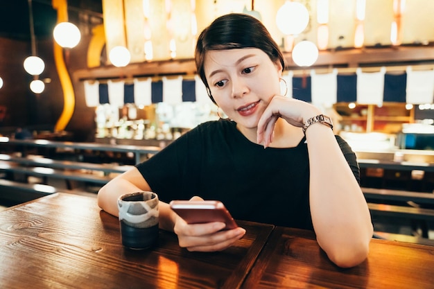 日本食レストランで食事を待っている間携帯電話でメッセージを読んでいる若いアジアの韓国人女性。熱い抹茶とピンクの携帯電話でウェブサイトで見ている美しいエレガントな女の子suferインターネット