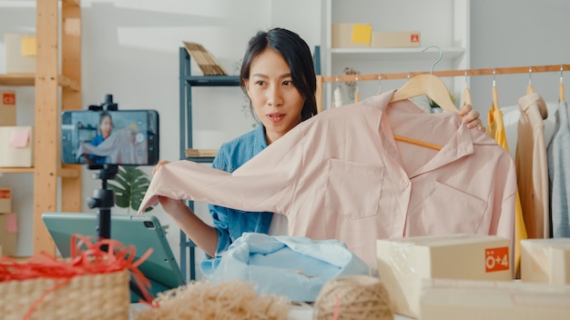 Young asian lady fashion designer using mobile phone receiving purchase order and show clothes in live streaming