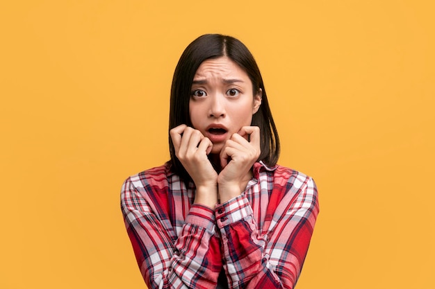 Young asian lady feeling scared touching cheeks with hands and looking at camera with open mouth yellow background